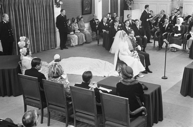 Schwarzes und weißes Foto einer Hochzeitszeremonie mit Menschen, die sitzen und stehen, einem Mann mit Mikrofon und einer Frau mit Blumenstrauß, sowie einem Podium und Blumenschmuck.
