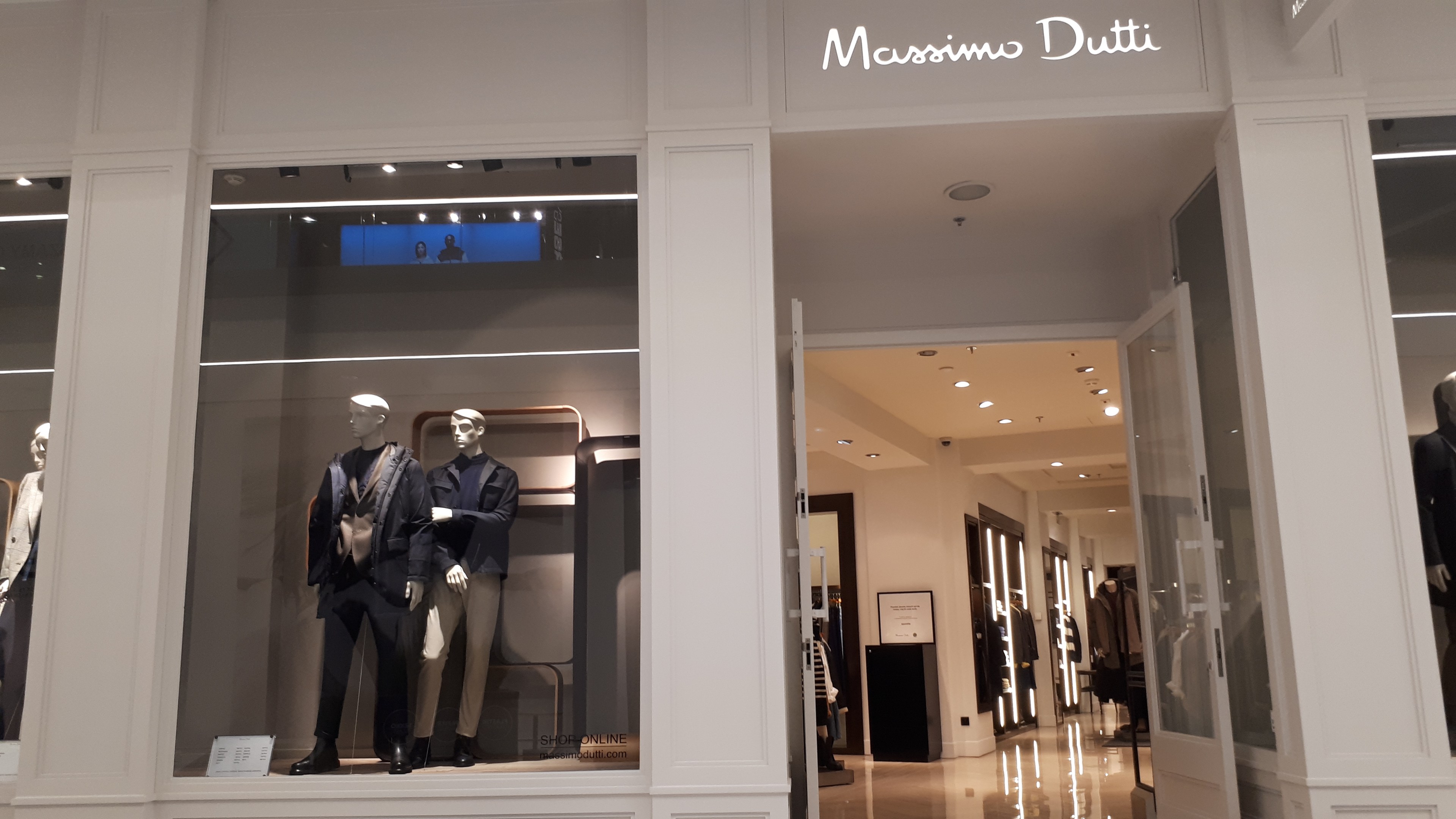 Vorderfront des Massimo Dutti in Mailand, Italien, mit Schaufensterpuppen in moderner Kleidung, beleuchteter Decke, Informationsschildern und sichtbarem Boden.