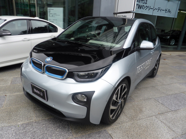 Ein silberner BMW i3 Elektroauto vor einem Gebäude in Tokyo, Japan, mit Glaswänden und einer Tafel im Hintergrund.