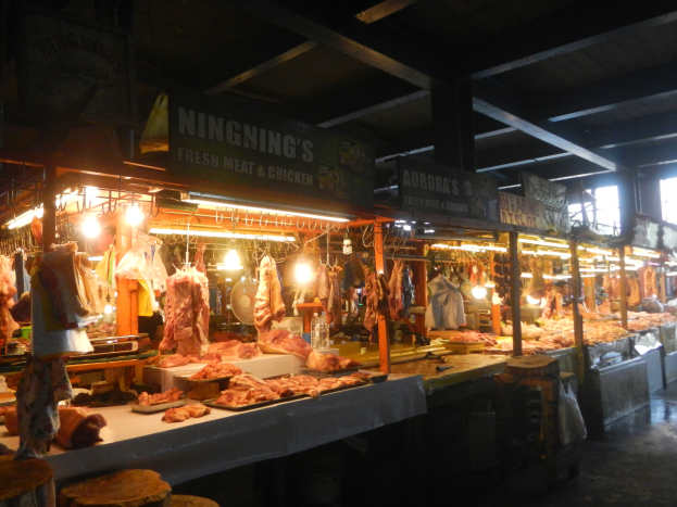 Ein Metzgerladen mit verschiedenen Fleischsorten auf Display, beleuchtet von Lampen, umgeben von Texttafeln, Hockern und anderen Gegenständen.