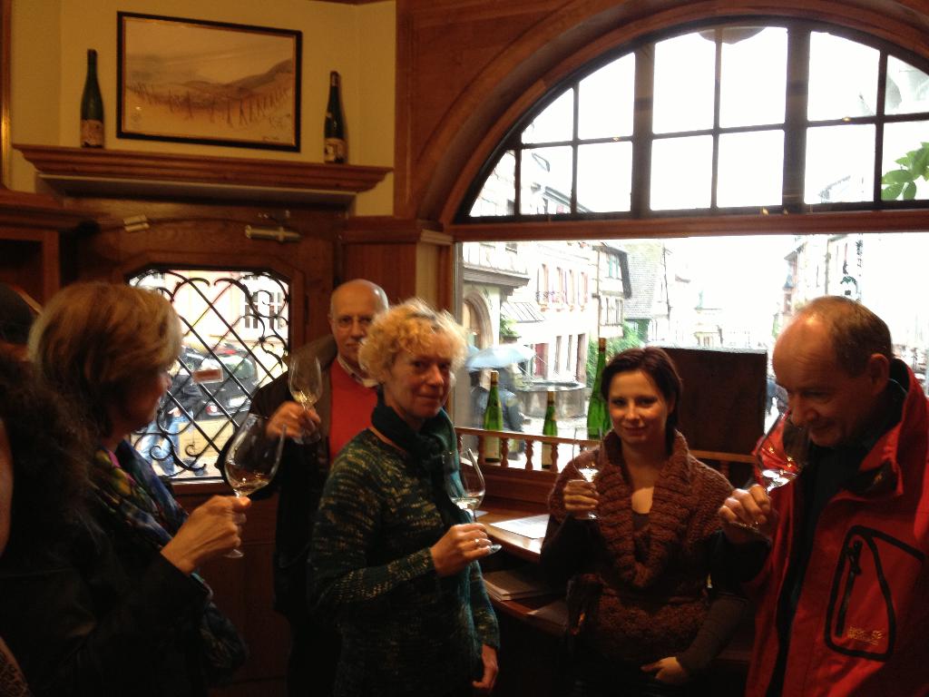 Fünf Personen mit Gläsern Alkohol vor einem Fenster oder einer Tür mit Flaschen im Hintergrund und weitere Gebäude im Hintergrund sichtbar, möglicherweise in einer Bar.