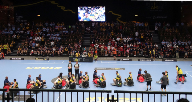 Eine Gruppe von Rollstuhlfahrern auf einem Platz bei den Invictus Games Düsseldorf 2020, mit einer großen Menge im Hintergrund und informativen Tafeln über den Platz verteilt.