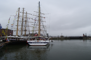 Mehrere Schiffe sind im Wasser mit Gebäuden, einer Fußgängerbrücke, Flaggen und Masten im Hintergrund unter einem bewölkten Himmel.