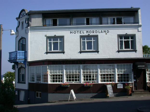 Das Hotel Nordland in Stavanger, Norwegen, ein mehrstöckiges Gebäude mit zahlreichen Fenstern, einem Schild mit Text, einem Straßenpfosten, einer Gruppe von Bäumen davor und einem bewölkten Himmel im Hintergrund.