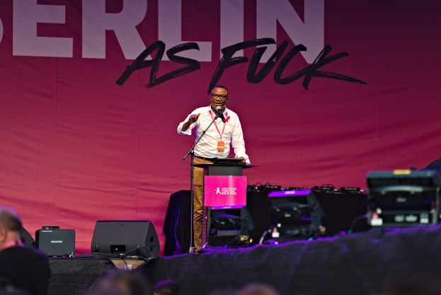 Ein Mann in einem weißen Hemd und braunen Hosen steht auf einer Bühne und hält ein Mikrofon in der Hand, während er eine Menge anspricht. Im Hintergrund ist ein pinkes Banner mit der Aufschrift "Berlin as Fuck" zu sehen.