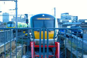 Zwei Züge auf parallelen Gleisen mit einem Zaun im Vordergrund, ein Auto auf der Straße rechts, eine Signalleuchte auf einem Pfahl, Gebäude im Hintergrund und der Himmel darüber.