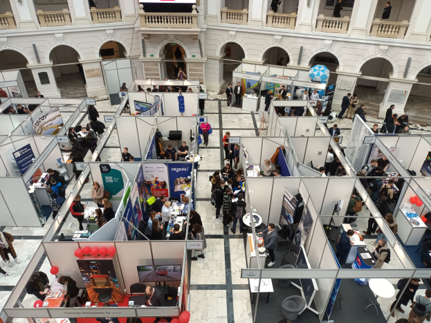 Große Besucherzahl auf einer internationalen Fachmesse mit Ständen, Bannern, Tischen, Stühlen, Ballons und einem Gebäude mit Bögen, Säulen, Geländern und einem Bildschirm im Hintergrund.
