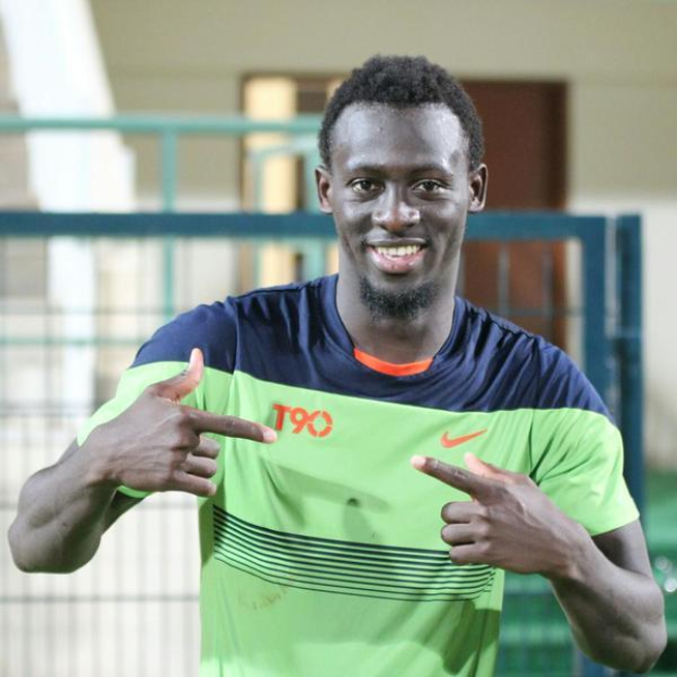 Ein Mann in einem grün-blauen Hemd, identifiziert als Emmanuel Adebayor, zeigt mit dem Finger auf die Kamera und lächelt, mit einem unscharfen Geländer, einer Wand und einer Tür im Hintergrund.