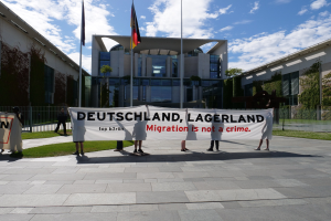 Eine Gruppe von Menschen hält ein Transparent mit der Aufschrift "Deutschland, Lagerland Migration ist kein Verbrechen" vor einem Gebäude mit Fenstern, umgeben von Gras, einem Metallzaun, Pflanzen, Bäumen und Fahnenstangen.