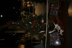 Ein geschmückter Weihnachtsbaum mit Geschenken, Rahmen und Spielzeug auf einer Plattform, mit Menschen, zusätzlichen Weihnachtsbäumen und Lichtern im Hintergrund.