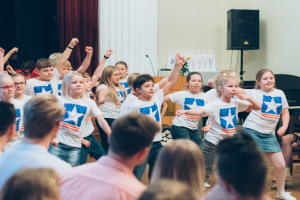Eine Gruppe von Kindern in weißen Hemden und blauen Shorts tanzt vor einem sitzenden Publikum, mit einer Vorhangwand, Lautsprecherständer und einem Tisch mit Mikrofon im Hintergrund.