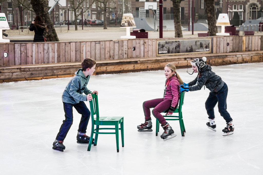 Kinder auf Skiern im Vordergrund eines Spielplatzes, mit drei Kindern und zwei Stühlen in der Mitte und Gebäuden, Bäumen, Bänken, Pfosten und einem Basketballfeld im Hintergrund.