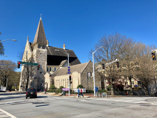 Große Kirche mit Turm an der Ecke einer Straße, identifiziert als St. Lukes Episcopal Church, umgeben von Gebäuden, Straßenmöbeln, Fahrzeugen, Passanten, Grünflächen und einem klaren Himmel.