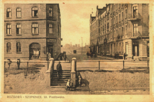 Ein altes Foto einer Stadtstraße mit Gebäuden, Menschen, Treppen, Pfählen und einem Zaun, mit Text am unteren Bildrand.