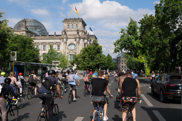 Eine Gruppe von Menschen, die Fahrräder auf einer von Bäumen gesäumten Straße in Berlin, Deutschland, fahren, mit einer Bushaltestelle auf der rechten Seite und einer Flagge, die auf einem Gebäude unter einem bewölkten Himmel weht.