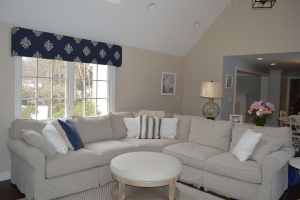 Ein Wohnzimmer mit einer Couch, Kissen, einem Tisch, Stühlen, einem Blumenstrauß, einem Foto Rahmen, einer Alarm Uhr und einem Fenster auf der linken Seite mit an der Decke angebrachten Lampen.