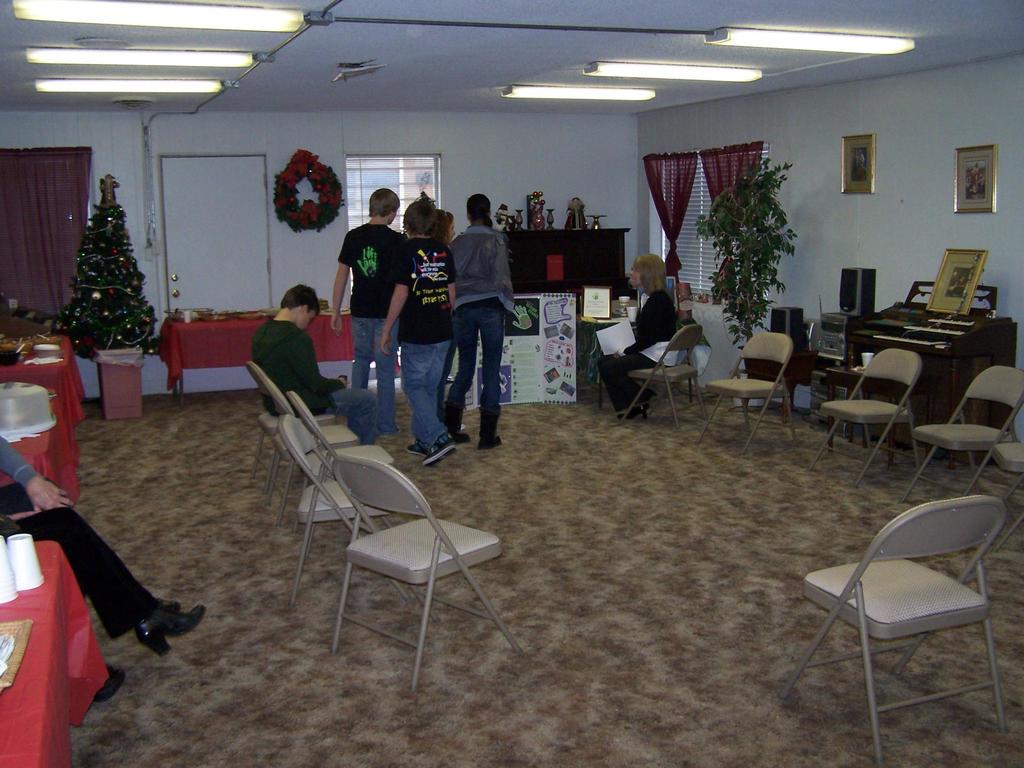 Ein Raum mit mehreren Stühlen und Tischen, einige Personen stehen und andere sitzen, Tassen und Gefäße auf den Tischen, eine Wand mit einem Strauß, Vorhang, Tür, Foto Rahmen und Fenstern, ein Weihnachtsbaum, ein Piano auf der rechten Seite und Röhrenleuchten an der Decke.