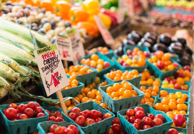 Ein Bauernmarkt mit Körben voller frischer Erzeugnisse, darunter Tomaten und Mais, sowie Schilder mit Text im Hintergrund.