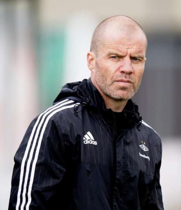 Ein kahlköpfiger Mann in einer schwarzen Jacke steht auf einem Fussballfeld, mit einer marineblauen Jacke mit weissen Streifen und einem weissen Kragen, mit verschränkten Händen und einem ernsten Gesichtsausdruck.