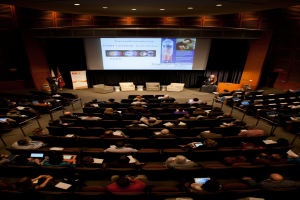 Ein vollbesetzter Hörsaal mit sitzenden Menschen, die Gegenstände halten, beleuchtet von vielen Lichtern, mit Fahnen links, einem Podium rechts und einer Werbetafel links.