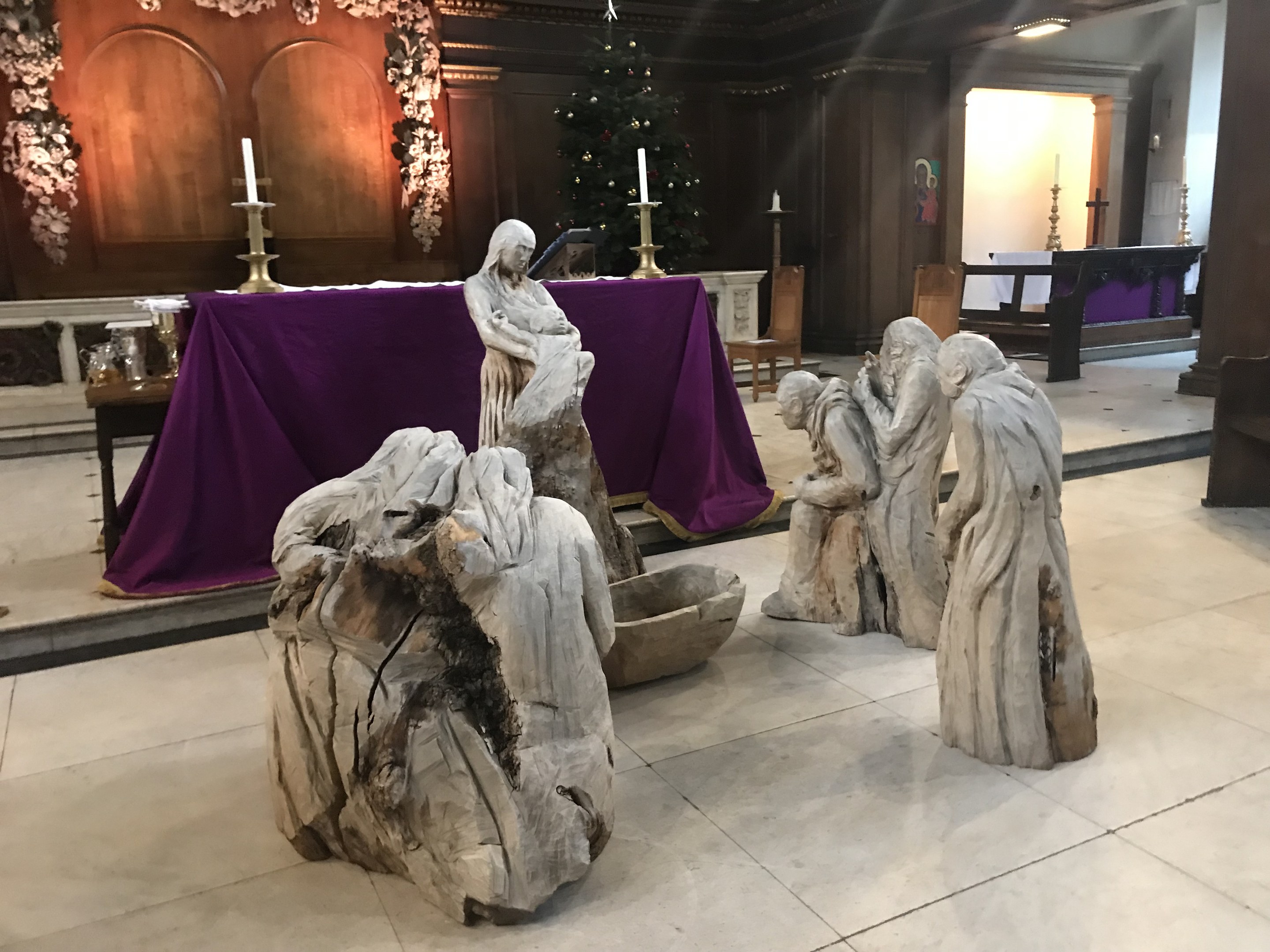 Krippe in einer Kirche mit Statuen auf dem Boden, einem Tisch mit einer Decke und Kerzenständern, Stühlen, einem geschmückten Weihnachtsbaum und einer Wand im Hintergrund.