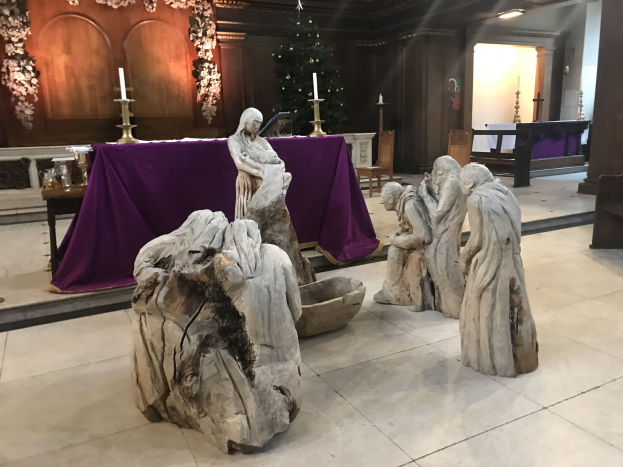 Krippe in einer Kirche mit Statuen auf dem Boden, einem Tisch mit einer Decke und Kerzenständern, Stühlen, einem geschmückten Weihnachtsbaum und einer Wand im Hintergrund.