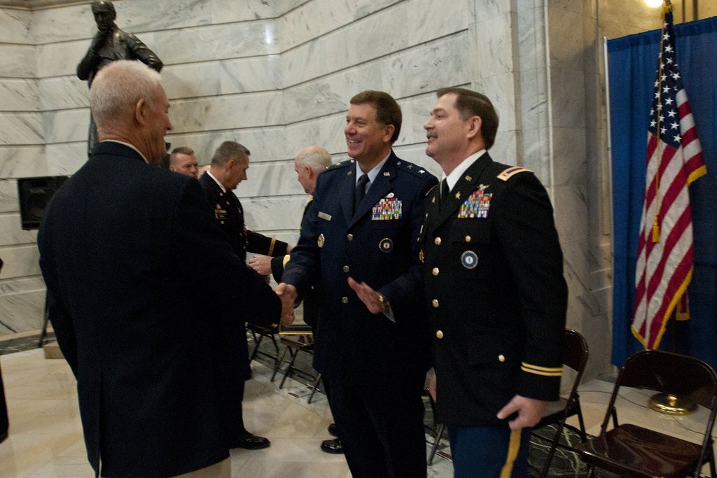 Menschen schütteln sich die Hände vor Stühlen, mit einer Statue, einer Flagge und einem Vorhang im Hintergrund.