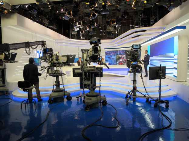 Ein TV-Studio mit mehreren Kameras auf dem Boden, einer sitzenden Person im Vordergrund und einigen stehenden Personen im Hintergrund, mit an der Decke montierten Lichtern für eine Live-Broadcast-Aufnahme.