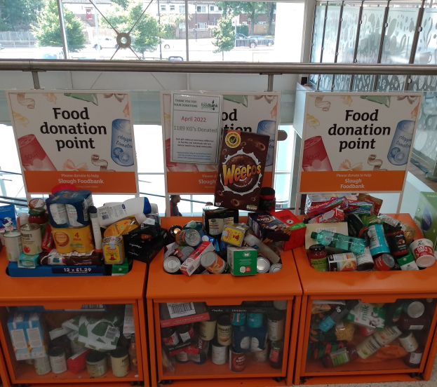 Orangene Schübe mit Lebensmittelspenden, darunter Flaschen, Dosen und Kisten, mit Informationsschildern in der Nähe, durch Glasfenster mit Außenfahrzeugen, Bäumen, Gebäuden und Himmel.