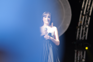 Eine Frau in einem blauen Kleid steht selbstbewusst auf einer hell erleuchteten Bühne und hält ein Mikrofon in der Hand, während das Publikum im Hintergrund zu sehen ist.