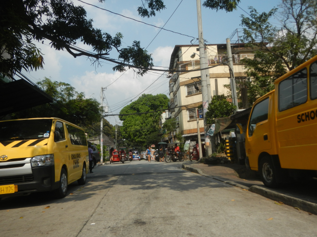 Ein gelber Schulbus fährt auf einer Straße mit hohen Gebäuden, Bäumen und Strommasten mit Drähten, mit Menschen auf Fahrrädern und anderen Fahrzeugen auf der Straße, unter einem bewölkten Himmel.