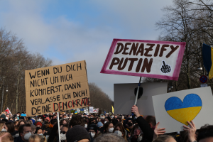 Eine große Gruppe von Menschen mit Schildern "Denazifiziere Putin" und Flaggen, die vor einer Menge mit Bäumen und einem klaren Himmel demonstrieren.