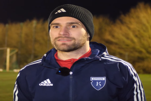 Ein Mann in einer blauen Jacke und Mütze steht auf einem Fußballfeld, umgeben von Bäumen, mit einem Tor im Hintergrund, während eines Trainings.