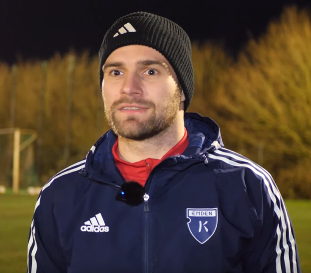 Ein Mann in einer blauen Jacke und Mütze steht auf einem Fußballfeld, umgeben von Bäumen, mit einem Tor im Hintergrund, während eines Trainings.