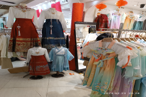 Ein Geschäft mit Schaufensterpuppen in traditioneller koreanischer Hanbok-Kleidung, Kleider an Bügeln, eine Kartonschachtel auf dem Boden, ein Spiegel, eine Säule und eine Decke mit Lampen.