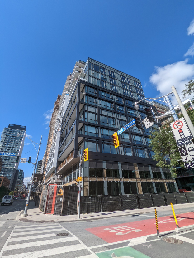 Eine belebte Stadtstraße mit einem hohen Apartment/Wohnhausgebäude zum Verkauf an der 541 East 57th Street, Unit 541, Fahrzeugen, Verkehrsampeln, Schildern, Bäumen, einem Zaun und einem bewölkten Himmel.