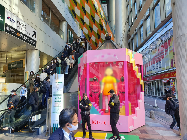 Eine Gruppe von Menschen steht vor einem Gebäude mit Glaswänden, Texttafeln, einige tragen Masken, in der Nähe einer Rolltreppe; ein pinkfarbener Kasten in der Mitte zeigt Schaufensterpuppen und trägt die Aufschrift "Netflix's new Netflix booth in Tokyo."