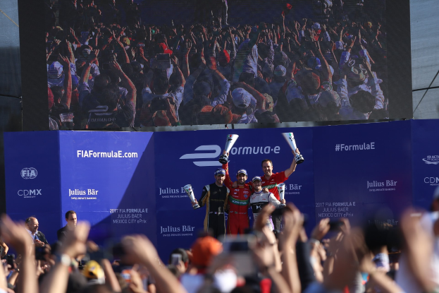 Podium mit einer Gruppe von Menschen mit Pokale, beschriftet mit "FIA Formula E Mexico City", einem Bildschirm und Himmel im Hintergrund.