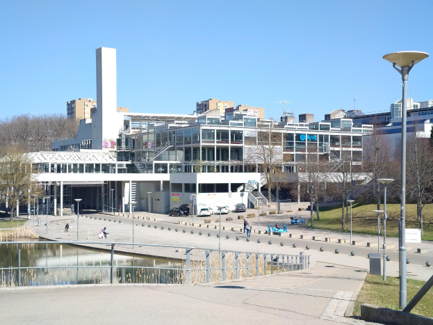 Großes Gebäude mit einem Teich davor, umgeben von Straßenmasten, Straßenlaternen, Fahrzeugen, Fußgängern, Absperrpollern, Bäumen und einem bewölkten Himmel, identifiziert als der neue Campus der Universität Toronto.