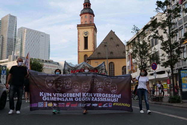 Maskierte Menschen tragen ein Transparent die Straße entlang an einem geparkten Auto vorbei, mit Gebäuden, Bäumen, Schildern, Pfosten, einem Uhrenturm und einem klaren blauen Himmel im Hintergrund.