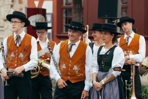 Gruppe von Menschen in traditioneller bayrischer Tracht mit Musikinstrumenten, vor einem Gebäude mit Fenstern, einem Lautsprecher und Pflanzen im Hintergrund.