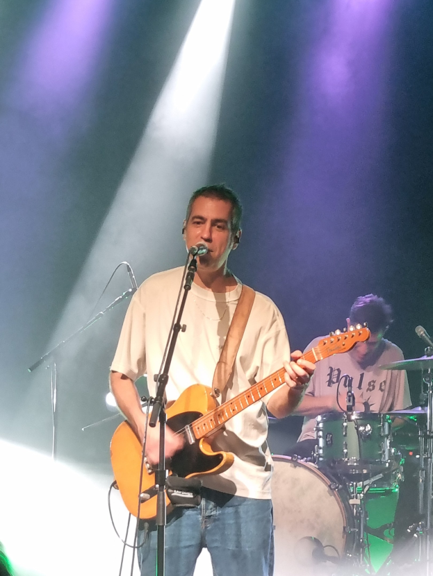 Ein Mann spielt Gitarre und singt in ein Mikrofon auf der Bühne, während ein anderer Mann Schlagzeug spielt.