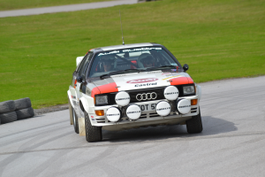 Ein Audi Quattro S1 Rallyeauto fährt mit zwei Helmträgern darin auf der Straße, Reifen sind auf der linken Seite und Gras ist im Hintergrund zu sehen.