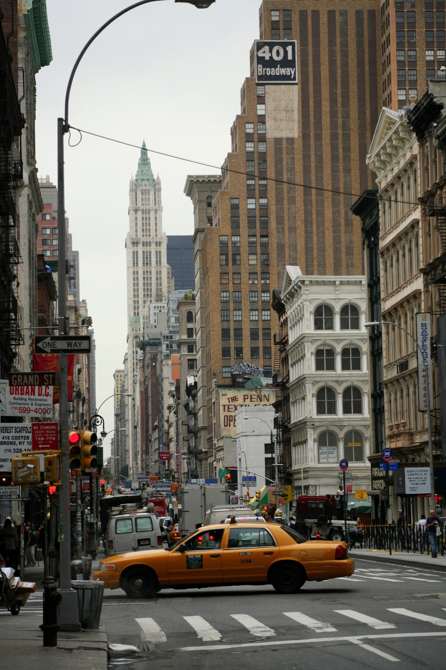 Gelbes Taxi fährt auf einer belebten Stadtstraße mit hohen Gebäuden, Passanten auf dem Gehweg, Ampeln und Textanzeigen, unter einem sichtbaren Himmel.