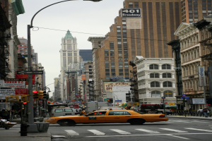 Gelbes Taxi fährt auf einer belebten Stadtstraße mit hohen Gebäuden, Passanten auf dem Gehweg, Ampeln und Textanzeigen, unter einem sichtbaren Himmel.