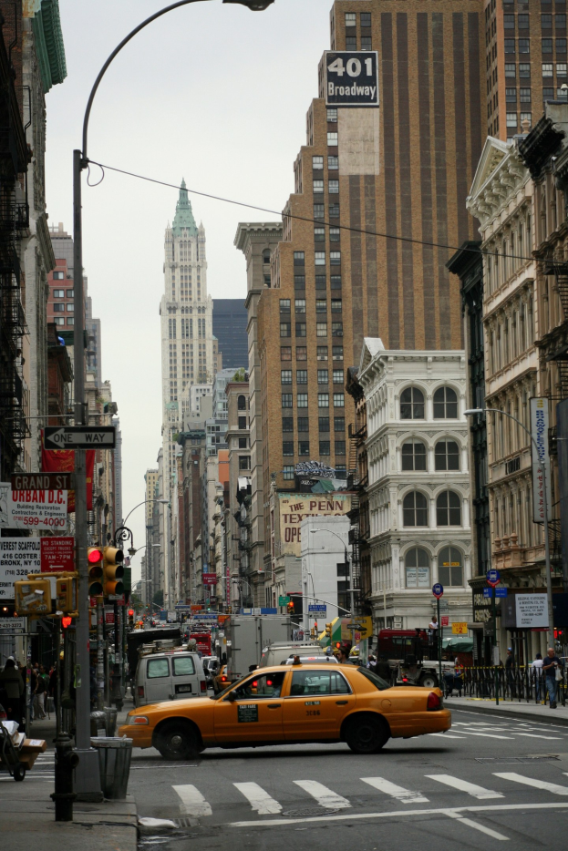 Gelbes Taxi fährt auf einer belebten Stadtstraße mit hohen Gebäuden, Passanten auf dem Gehweg, Ampeln und Textanzeigen, unter einem sichtbaren Himmel.