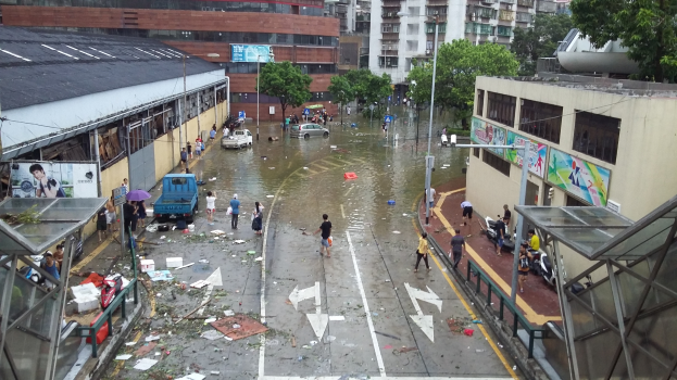 Eine überflutete Stadtstraße mit Menschen, einige mit Schirmen, umgeben von Gebäuden, Bäumen, Laternenmasten und Fahrzeugen, mit verstreuten Trümmern und einer Hütte auf der rechten Seite.