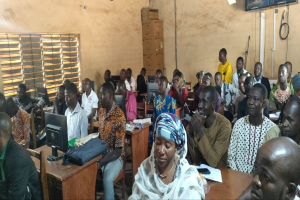 Eine Gruppe von Menschen sitzt an Tischen in einem classroom mit Monitoren, Taschen, Büchern und Papieren auf den Tischen, ein Fenster links und Kartons und ein Fernseher an der Wand im Hintergrund, an einer Techniktrainingssitzung teilnehmend.