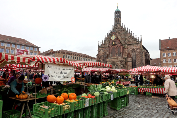 Ein belebter Markt in Nürnberg, Deutschland, mit farbenfrohen Obst- und Gemüseständen, Menschen mit Taschen und Zelten, vor Gebäuden mit Fenstern und einem Uhrenturm unter einem sichtbaren Himmel.