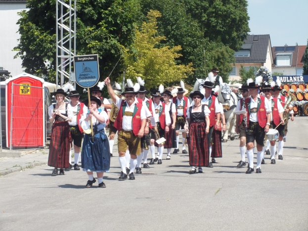 Eine Gruppe von Menschen in traditioneller bayrischer Tracht, die eine Straße entlanggehen, einige halten Musikinstrumente und tragen Hüte, mit Bäumen, Gebäuden und einem klaren blauen Himmel im Hintergrund und einem roten Objekt und einer Tafel mit Text auf der linken Seite.